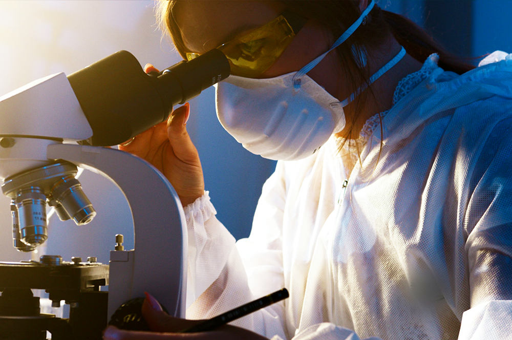 Mulher de óculos especiais, máscara e avental olha em um microscópio.